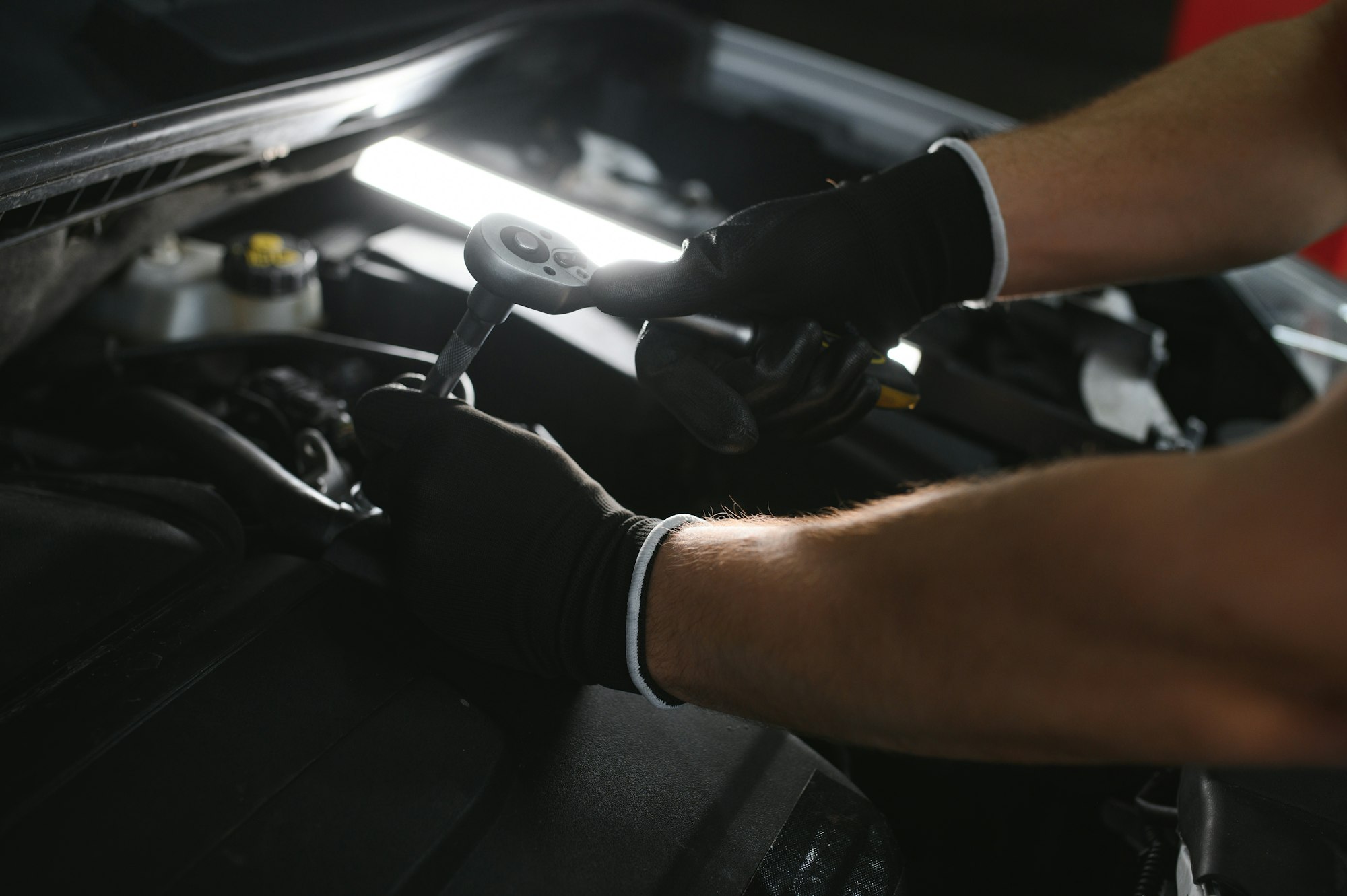 A mechanic is doing car checkup under the hood with flashlight at mechanic's shop.